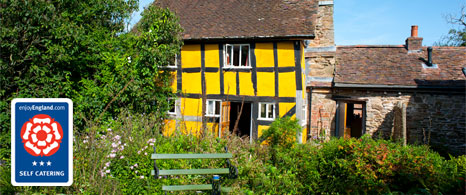 An exterior view of the historic yellow timber-framed cottage surrounded by lush greenery, overlaid with a VisitEngland 3-star self catering logo.